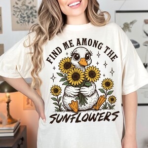 May include: White t-shirt with a graphic of a duck holding a bouquet of sunflowers. The text "Find me among the sunflowers" is printed on the shirt.