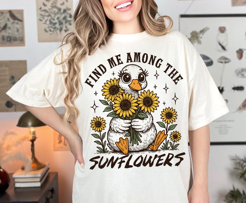 May include: White t-shirt with a graphic of a duck holding a bouquet of sunflowers. The text "Find me among the sunflowers" is printed on the shirt.