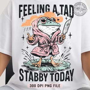 May include: White t-shirt featuring a cartoon frog wearing a pink robe and holding a knife. The frog is standing on a lily pad with the text "FEELING A TAD STABBY TODAY". The design includes a cloud, stars, and the text "300 DPI PNG FILE".