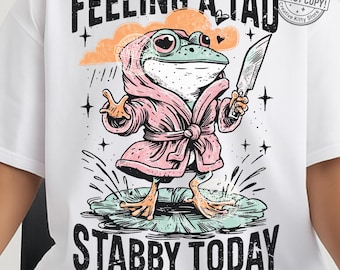 May include: White t-shirt featuring a cartoon frog wearing a pink robe and holding a knife. The frog is standing on a lily pad with the text "FEELING A TAD STABBY TODAY". The design includes a cloud, stars, and the text "300 DPI PNG FILE".