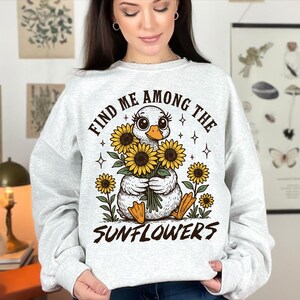 May include: A white sweatshirt with a graphic of a duck holding a bouquet of sunflowers. The text "Find me among the sunflowers" is printed on the sweatshirt.