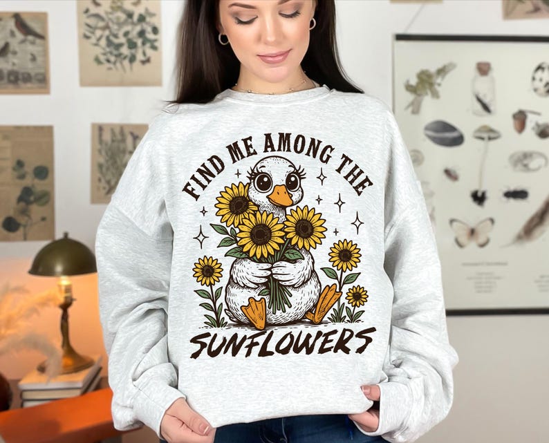 May include: A white sweatshirt with a graphic of a duck holding a bouquet of sunflowers. The text "Find me among the sunflowers" is printed on the sweatshirt.
