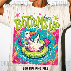 May include: Cream-colored t-shirt featuring a colorful graphic. The graphic includes the words "Bottoms Up" in green, a duck in a green inflatable ring, and confetti. The bottom of the shirt reads "300 DPI PNG FILE".