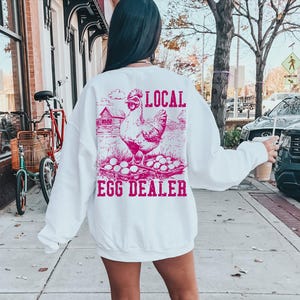 Op de afbeelding: Wit sweatshirt met een roze afbeelding van een kip en eieren. De tekst "LOCAL EGG DEALER" staat op het sweatshirt gedrukt.