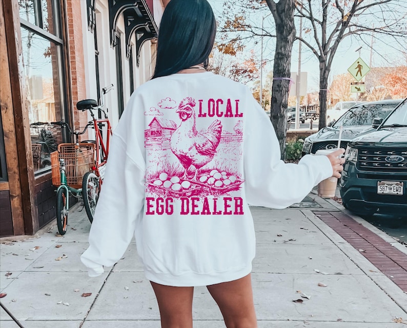 Op de afbeelding: Wit sweatshirt met een roze afbeelding van een kip en eieren. De tekst "LOCAL EGG DEALER" staat op het sweatshirt gedrukt.