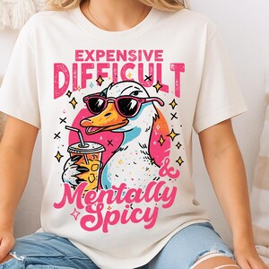Pu&ograve; includere: T-shirt color crema con un'anatra dei cartoni animati con occhiali da sole e una bevanda. La maglietta ha la scritta "Expensive, Difficult & Mentally Spicy" in rosa e bianco. L'anatra &egrave; bianca, gialla e blu.