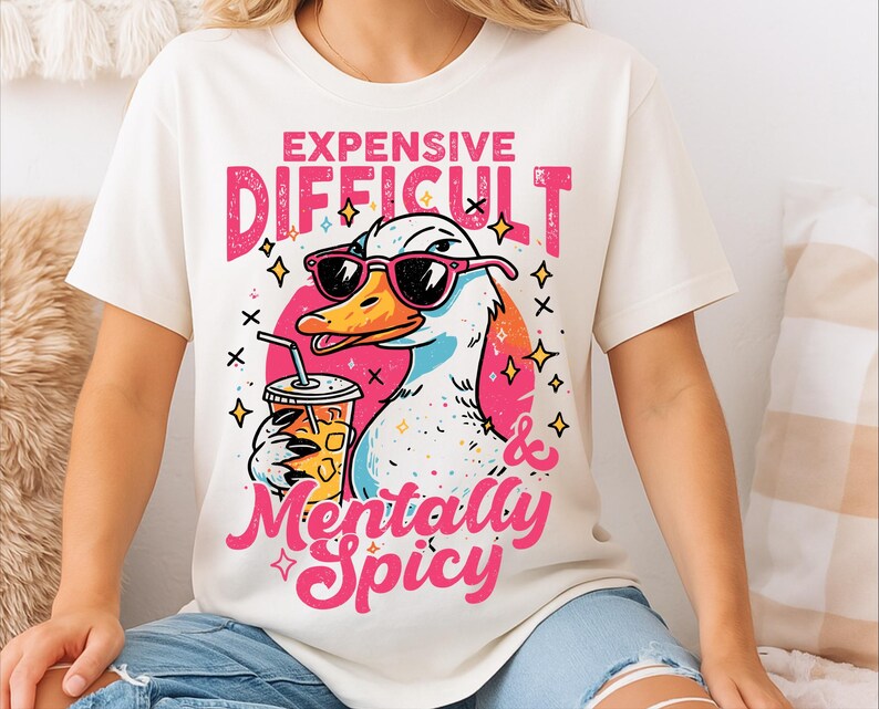 Pu&ograve; includere: T-shirt color crema con un'anatra dei cartoni animati con occhiali da sole e una bevanda. La maglietta ha la scritta "Expensive, Difficult & Mentally Spicy" in rosa e bianco. L'anatra &egrave; bianca, gialla e blu.