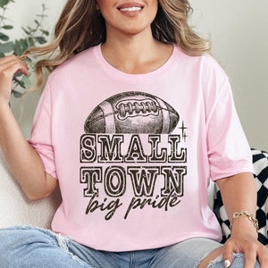 May include: A pink t-shirt with a vintage-style football graphic and the text "SMALL TOWN big pride".