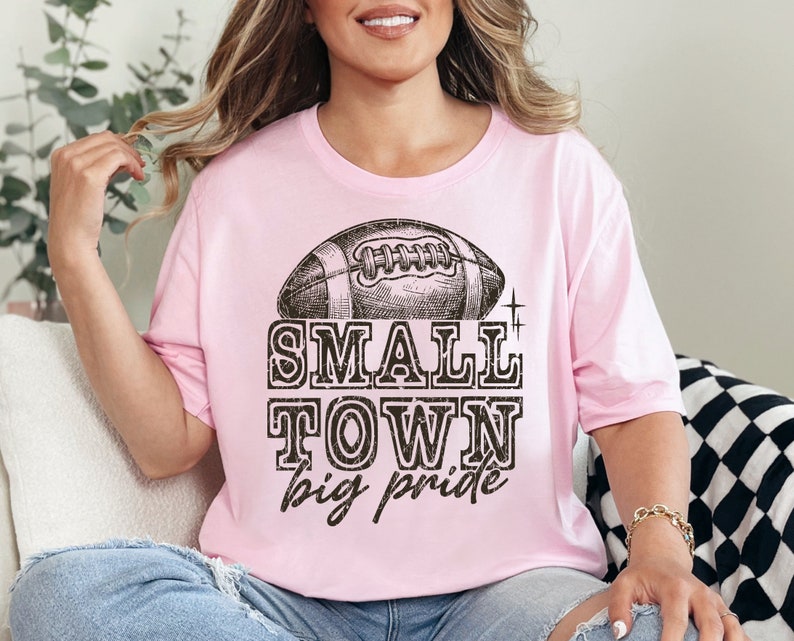 May include: A pink t-shirt with a vintage-style football graphic and the text "SMALL TOWN big pride".