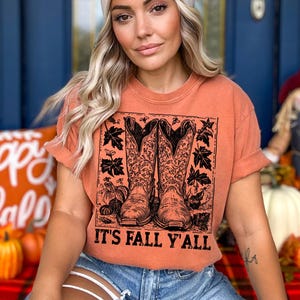 May include: Orange t-shirt featuring a graphic of cowboy boots, pumpkins, and leaves. The text "IT'S FALL Y'ALL" is printed below the graphic. The shirt is worn with blue denim shorts.