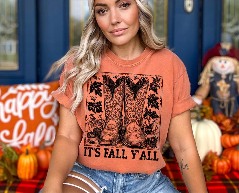 May include: Orange t-shirt featuring a graphic of cowboy boots, pumpkins, and leaves. The text "IT'S FALL Y'ALL" is printed below the graphic. The shirt is worn with blue denim shorts.