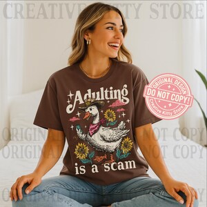 May include: A brown t-shirt featuring the phrase "Adulting is a scam" with a cartoon goose wearing a cowboy hat and bandana, set against sunflowers and pink clouds. The design includes stars.