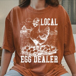 Op de afbeelding: Een roestkleurig T-shirt met een witte afbeelding van een kip en eieren. De tekst "LOCAL EGG DEALER" is op het shirt gedrukt.