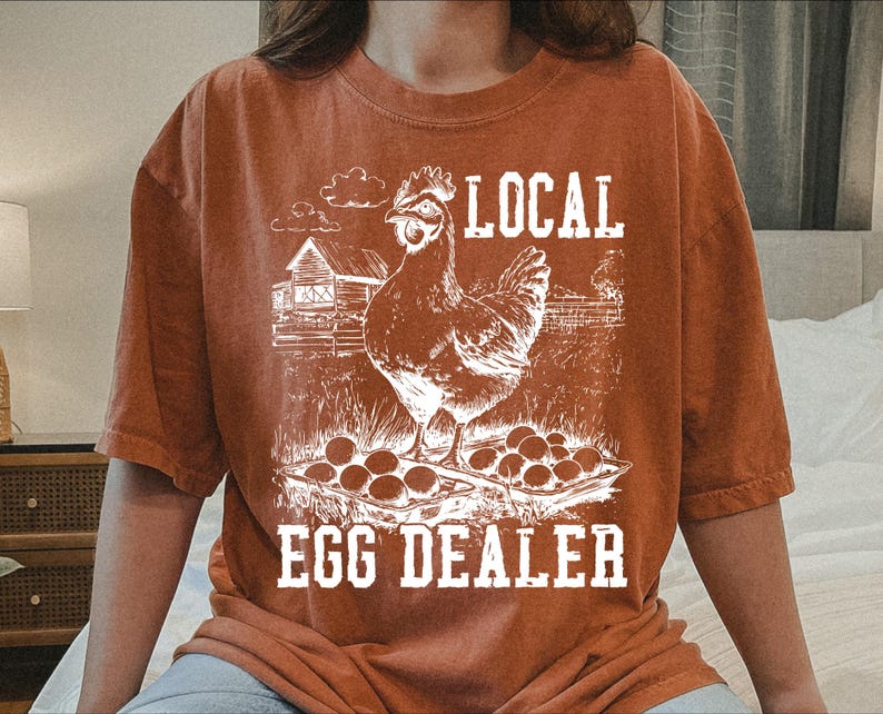Op de afbeelding: Een roestkleurig T-shirt met een witte afbeelding van een kip en eieren. De tekst "LOCAL EGG DEALER" is op het shirt gedrukt.
