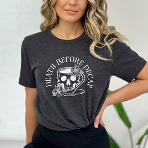 May include: Dark grey t-shirt with a white graphic. The graphic shows a skull in a teacup, roses, and the text "DEATH BEFORE DECAF". The shirt has short sleeves and a crew neck. The design is printed on the front.