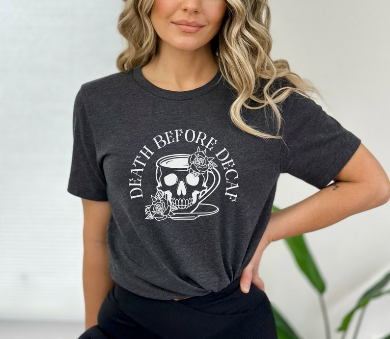May include: Dark grey t-shirt with a white graphic. The graphic shows a skull in a teacup, roses, and the text "DEATH BEFORE DECAF". The shirt has short sleeves and a crew neck. The design is printed on the front.