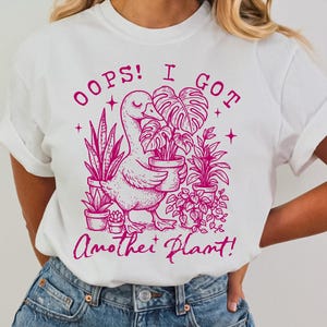 May include: White t-shirt with a pink graphic of a goose holding a potted plant. The text "Oops! I got another plant!" is printed on the shirt.