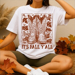 May include: White t-shirt featuring a brown graphic of cowboy boots, pumpkins, and autumn leaves. The text "IT'S FALL Y'ALL" is printed below the graphic. The shirt is worn by a person.