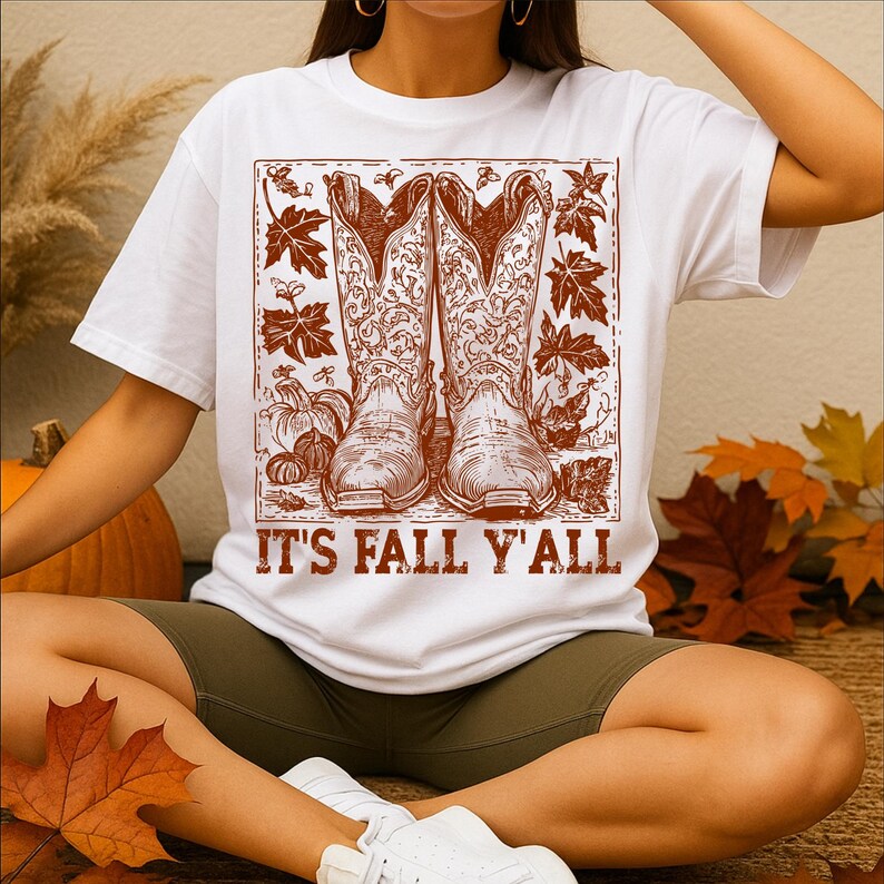 May include: White t-shirt featuring a brown graphic of cowboy boots, pumpkins, and autumn leaves. The text "IT'S FALL Y'ALL" is printed below the graphic. The shirt is worn by a person.