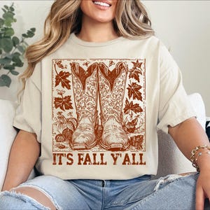 May include: A cream-coloured t-shirt with a brown graphic of cowboy boots, pumpkins, and autumn leaves. The text "IT'S FALL Y'ALL" is printed below. This casual t-shirt is a seasonal apparel item.