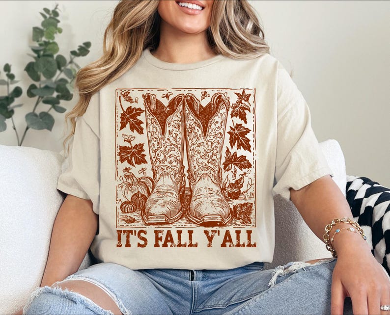 May include: A cream-coloured t-shirt with a brown graphic of cowboy boots, pumpkins, and autumn leaves. The text "IT'S FALL Y'ALL" is printed below. This casual t-shirt is a seasonal apparel item.