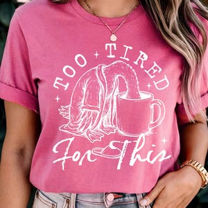 Op de afbeelding: Een roze t-shirt met een witte afbeelding van een cartoondier dat uit een mok drinkt. De tekst op het shirt luidt "Too Tired For This".