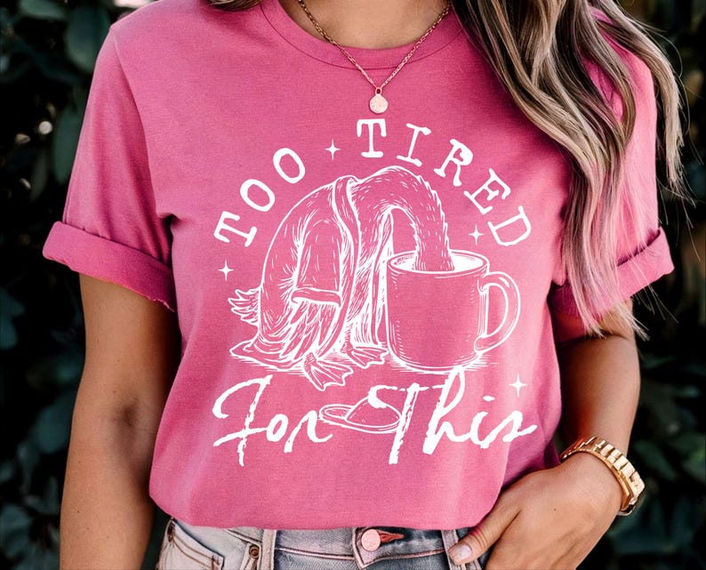 Op de afbeelding: Een roze t-shirt met een witte afbeelding van een cartoondier dat uit een mok drinkt. De tekst op het shirt luidt "Too Tired For This".