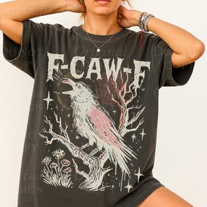 May include: Dark gray t-shirt with a distressed graphic featuring a crow perched on a branch, with the text "F-CAW-F". The crow has pink and white details, surrounded by stars and flowers. The shirt has a vintage, worn look.