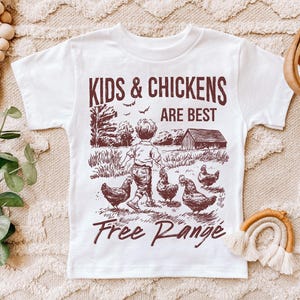 K&ouml;nnte beinhalten: Wei&szlig;es T-Shirt mit einer braunen Grafik eines Kindes, das mit H&uuml;hnern geht. Der Text auf dem Shirt lautet "Kids & Chickens Are Best Free Range".