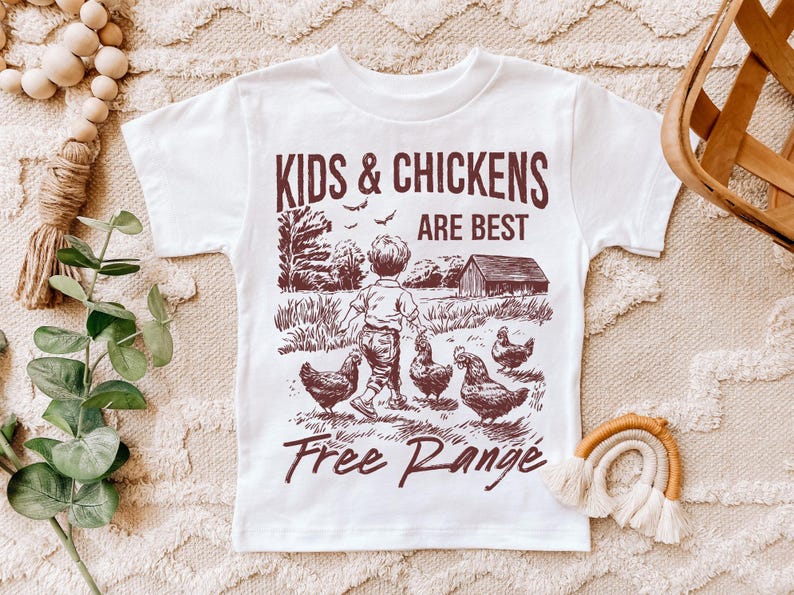 K&ouml;nnte beinhalten: Wei&szlig;es T-Shirt mit einer braunen Grafik eines Kindes, das mit H&uuml;hnern geht. Der Text auf dem Shirt lautet "Kids & Chickens Are Best Free Range".