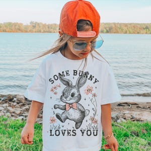 Peut inclure: Un t-shirt blanc avec une illustration en noir et blanc d'un lapin portant un n&oelig;ud papillon. Le texte "Some Bunny Loves You" est imprim&eacute; sur le t-shirt.