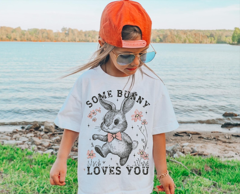 Peut inclure: Un t-shirt blanc avec une illustration en noir et blanc d'un lapin portant un n&oelig;ud papillon. Le texte "Some Bunny Loves You" est imprim&eacute; sur le t-shirt.