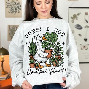 May include: A grey sweatshirt with a white goose holding a potted plant. The text on the sweatshirt reads "Oops! I got another plant!"