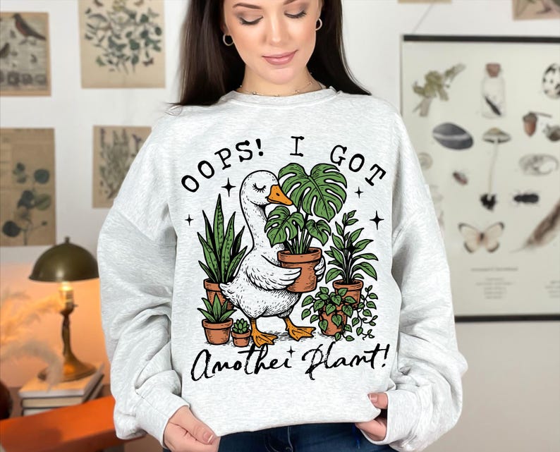 May include: A grey sweatshirt with a white goose holding a potted plant. The text on the sweatshirt reads "Oops! I got another plant!"