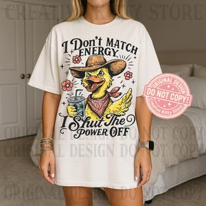 May include: Cream-colored t-shirt featuring a cartoon duck wearing a cowboy hat and bandana, holding a drink. The text reads "I Don't Match Energy, I Shut The Power Off." Pink floral accents and a "Do Not Copy" sticker are also visible.