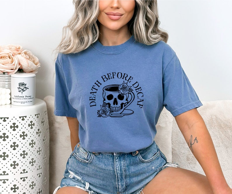 May include: A blue t-shirt with the phrase "DEATH BEFORE DECAF" in a circular design. The design features a skull inside a coffee cup with roses. The shirt is made of a soft material and is suitable for casual wear.