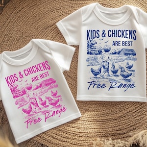 K&ouml;nnte beinhalten: Zwei wei&szlig;e T-Shirts mit einem blauen und rosa grafischen Aufdruck eines Kindes, das mit H&uuml;hnern geht. Der Text auf den Shirts lautet "Kids & Chickens Are Best Free Range".