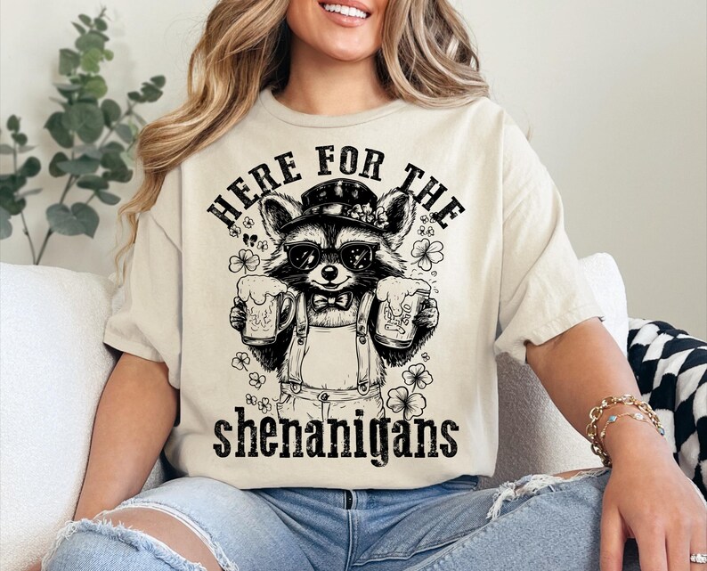 Puede incluir: Una camiseta beige con un gr&aacute;fico negro de un mapache con un sombrero y sosteniendo dos tazas. El texto "Here for the shenanigans" est&aacute; impreso debajo del mapache.