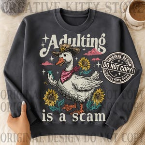 May include: Dark grey sweatshirt featuring a cartoon goose in a cowboy hat and bandana, holding a phone. The text reads "Adulting is a scam" with sunflowers and clouds. Includes a "Original Design Do Not Copy" sticker.