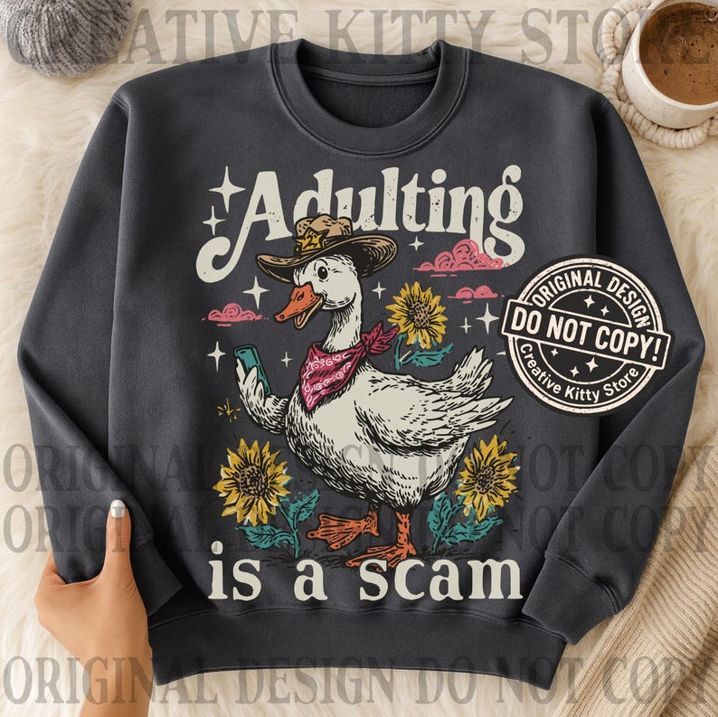 May include: Dark grey sweatshirt featuring a cartoon goose in a cowboy hat and bandana, holding a phone. The text reads "Adulting is a scam" with sunflowers and clouds. Includes a "Original Design Do Not Copy" sticker.