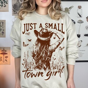 May include: A beige sweatshirt with the text "Just a small town girl" printed in brown. The graphic features a woman wearing a cowboy hat and a brown dress.