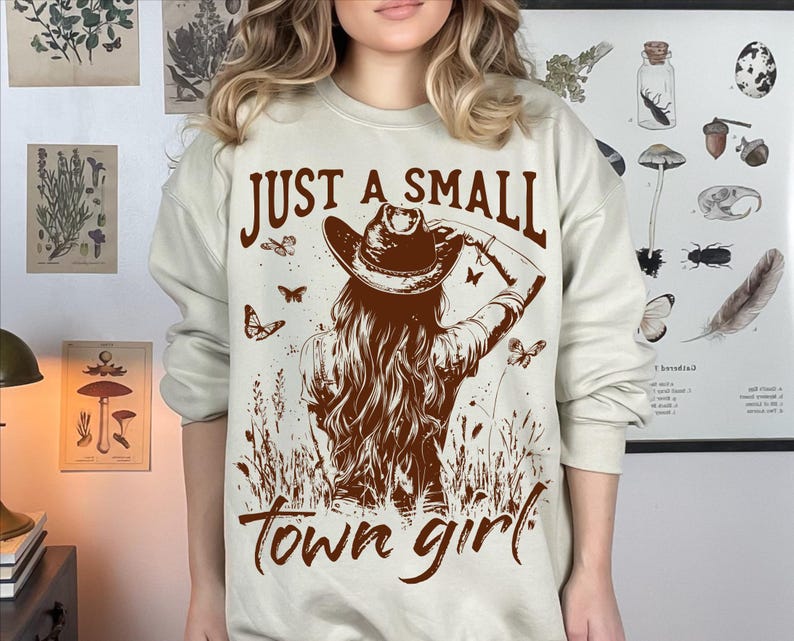 May include: A beige sweatshirt with the text "Just a small town girl" printed in brown. The graphic features a woman wearing a cowboy hat and a brown dress.