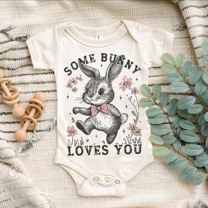 Peut inclure: Un body b&eacute;b&eacute; blanc avec une illustration en noir et blanc d'un lapin avec des fleurs roses et le texte "Some Bunny Loves You".