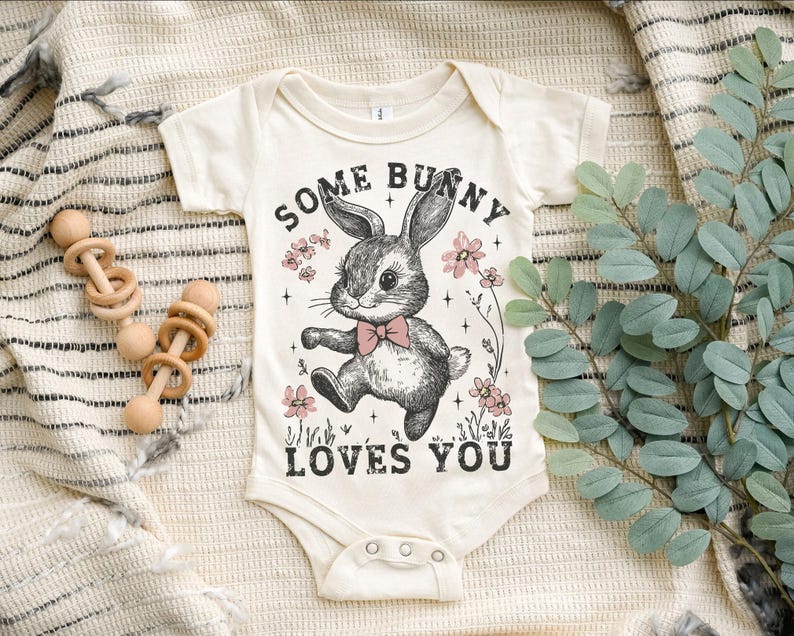 Peut inclure: Un body b&eacute;b&eacute; blanc avec une illustration en noir et blanc d'un lapin avec des fleurs roses et le texte "Some Bunny Loves You".