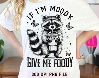 May include: White t-shirt with a black and white illustration of a raccoon holding a cup of coffee. The text "If I'm Moody Give Me Foody" is printed on the shirt.
