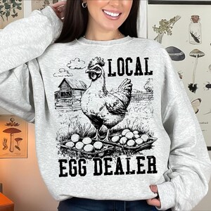 Op de afbeelding: Een grijs sweatshirt met een zwart-witte illustratie van een kip en de tekst "LOCAL EGG DEALER".
