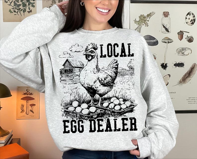 Op de afbeelding: Een grijs sweatshirt met een zwart-witte illustratie van een kip en de tekst "LOCAL EGG DEALER".