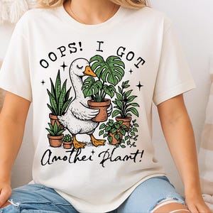 May include: White t-shirt featuring a graphic of a goose carrying a potted plant. The text "Oops! I got another plant!" is printed below the image.