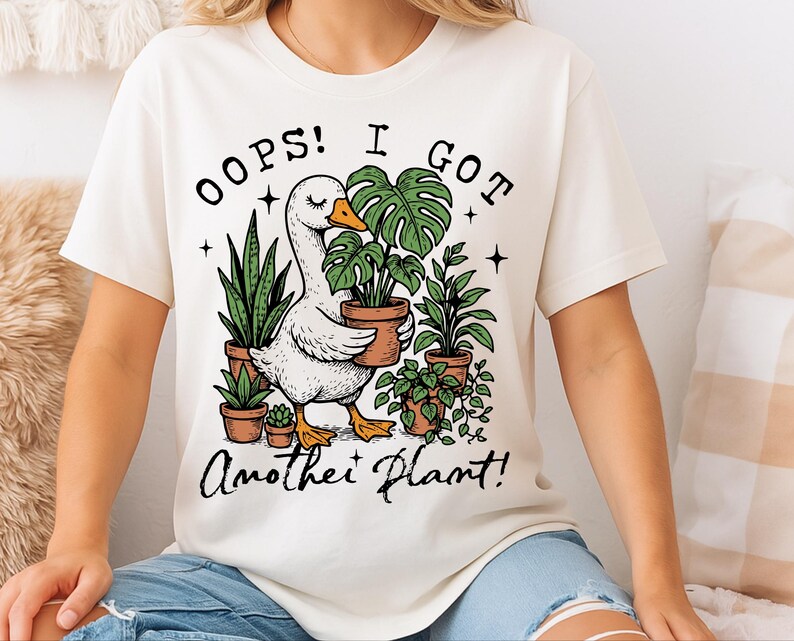May include: White t-shirt featuring a graphic of a goose carrying a potted plant. The text "Oops! I got another plant!" is printed below the image.