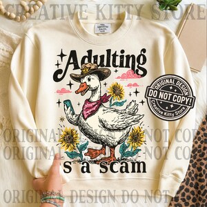 May include: Cream sweatshirt featuring a cartoon goose in a cowboy hat and bandana, holding a phone. The text "Adulting's a scam" is displayed with sunflowers and clouds. A Creative Kitty Store design.